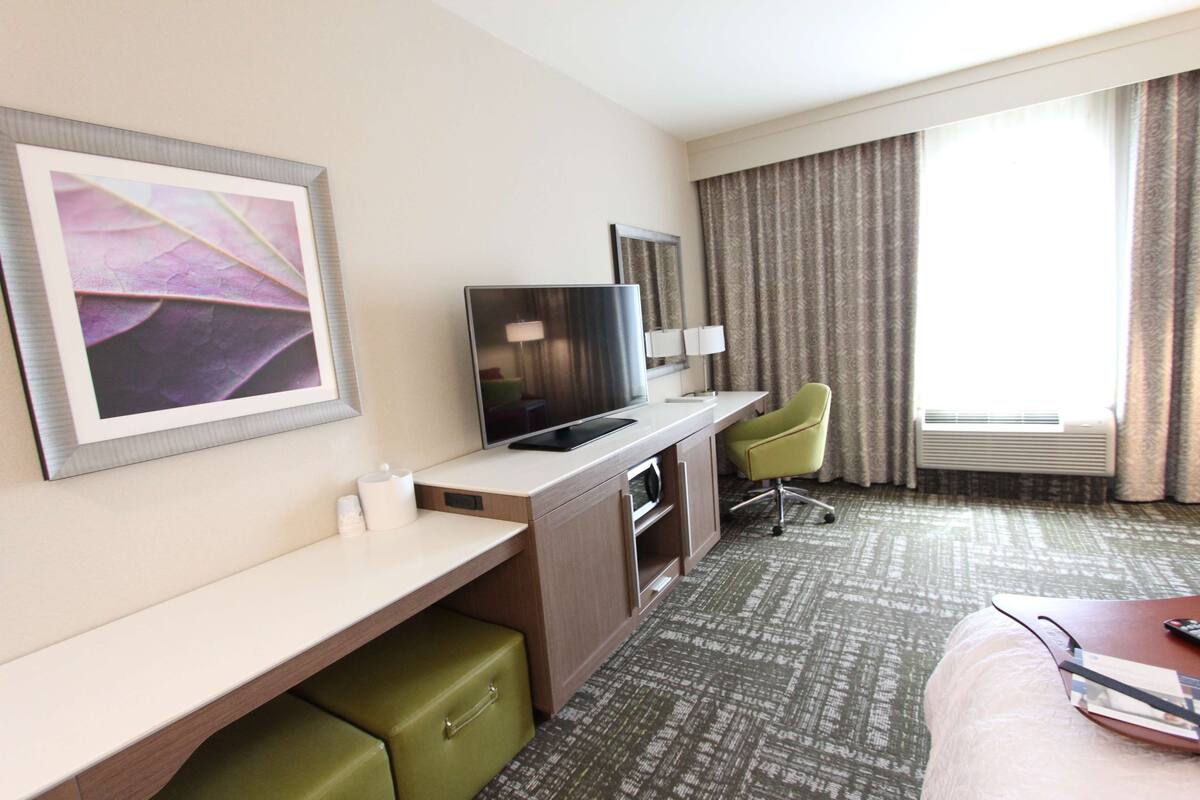 A hotel room with a bed, a desk with a television, a chair, and a large window with curtains.