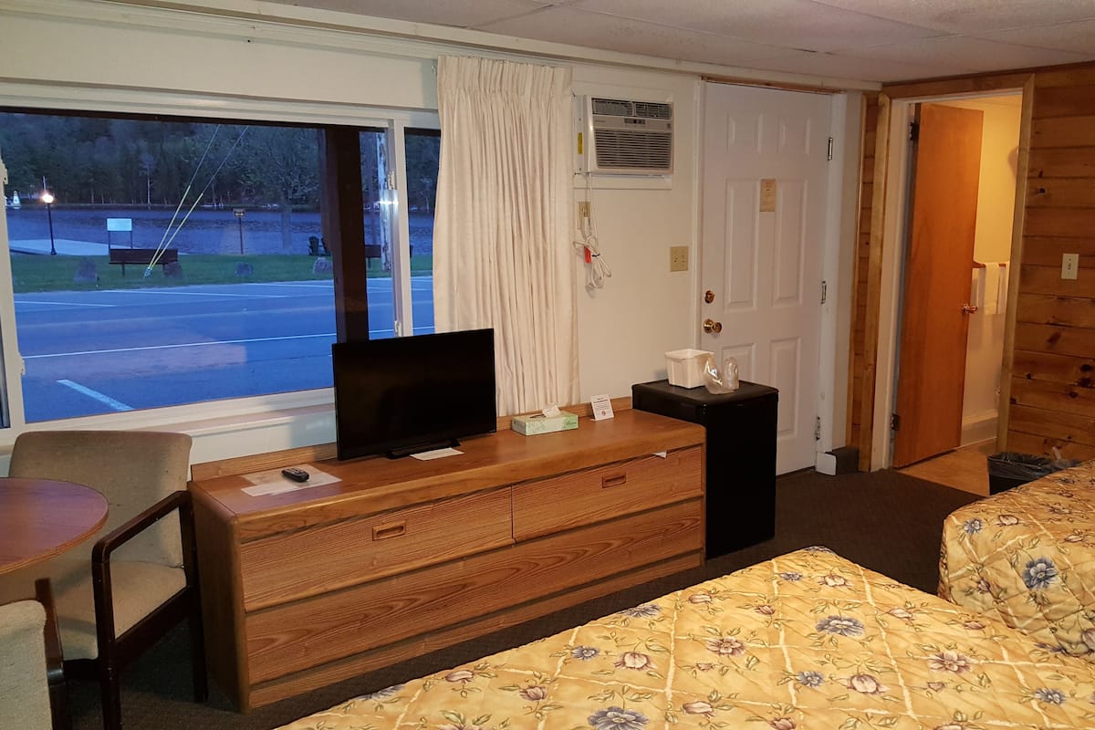 Quarto de hotel com cômoda de madeira, televisão em cima, cama com roupa de cama florida, janela com vista para um estacionamento e porta que dá para outro quarto.