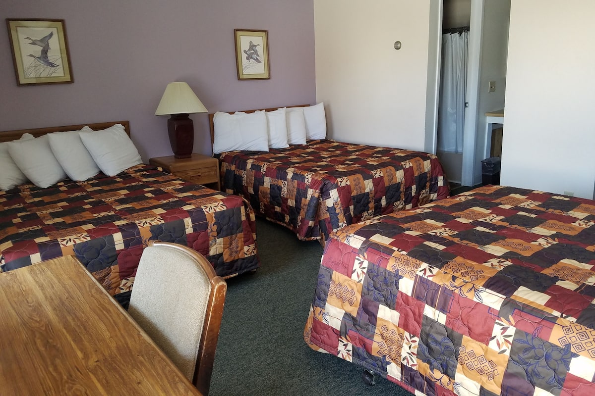 Quarto de hotel com duas camas, uma escrivaninha de madeira, mesa de cabeceira com abajur e porta de acesso a um banheiro.