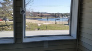 A window with a 'No Smoking' sign, a view of a lake, a building across the water, and a fenced area with a lamp post. - Clark's Beach Motel (Old Forge)