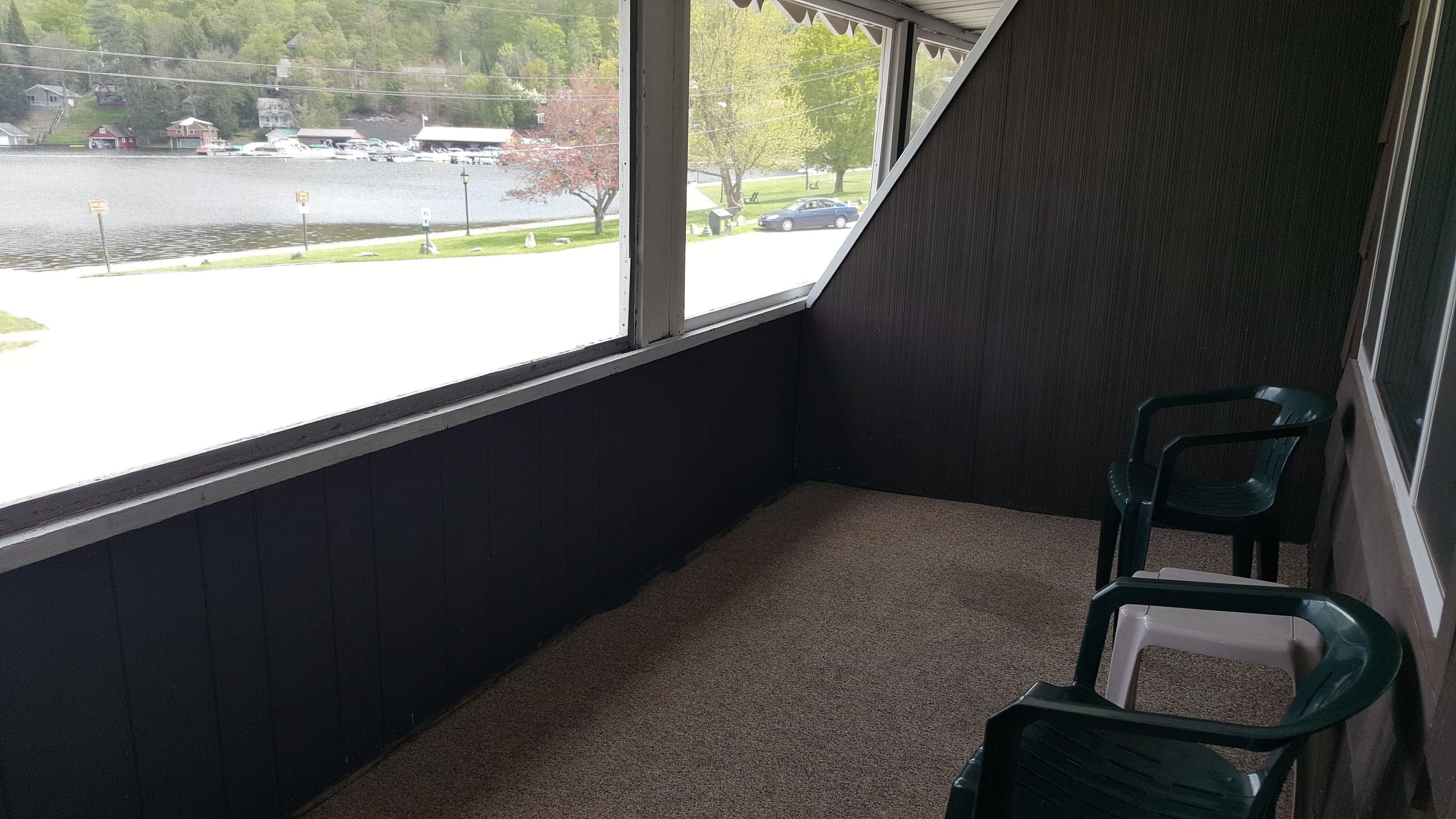 A corner room with a window overlooking a parking lot and a body of water. There are two green plastic chairs and a white trash bin.