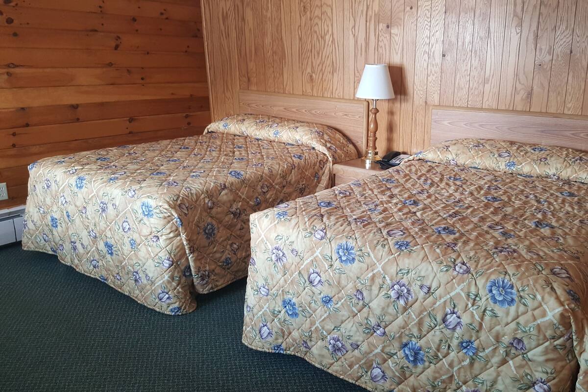 Duas camas de solteiro com roupa de cama florida, uma cabeceira de madeira, um abajur e um telefone numa parede de madeira.