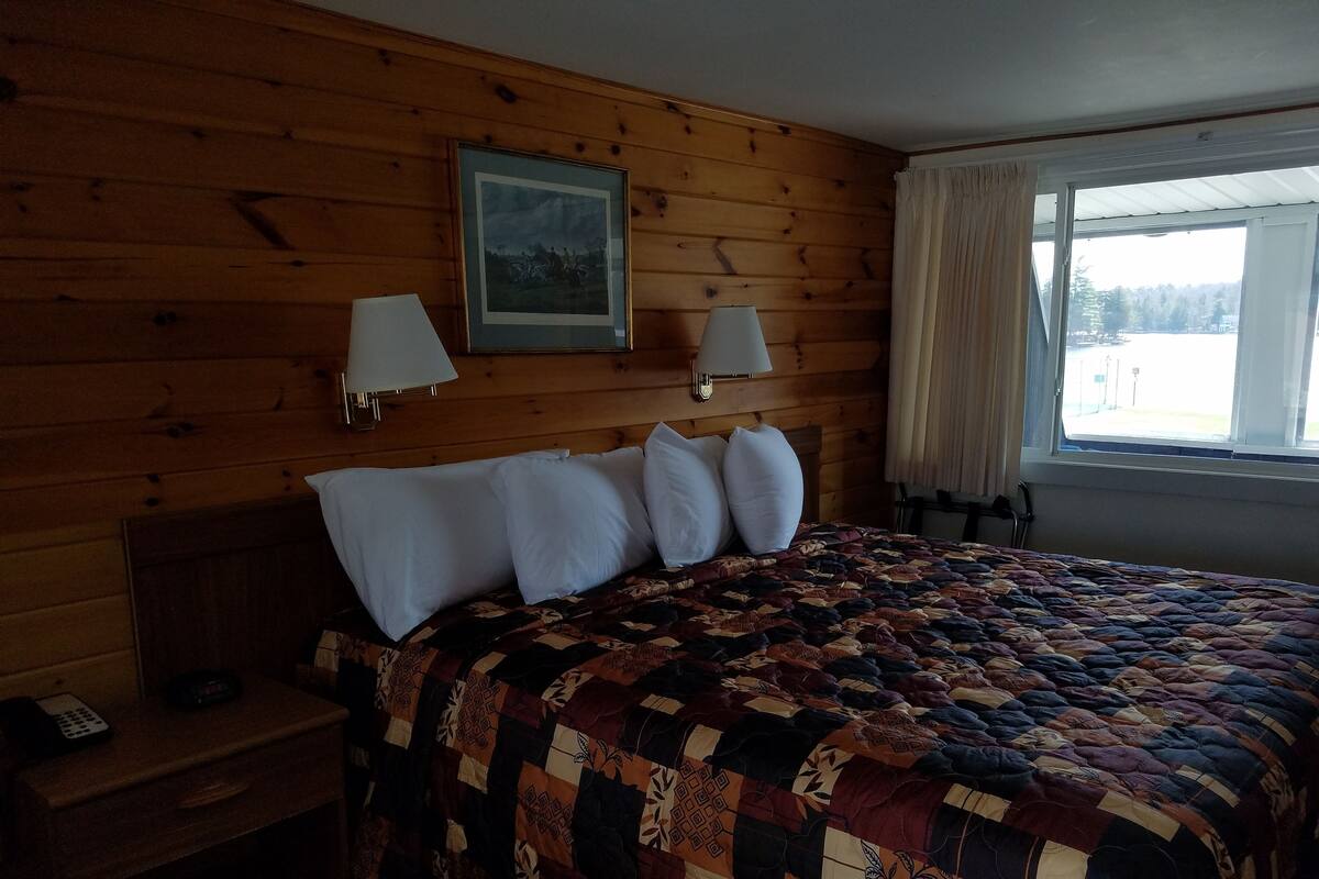 Um quarto com painéis de madeira, uma cama, duas luminárias na parede, um quadro emoldurado e uma janela com cortinas.