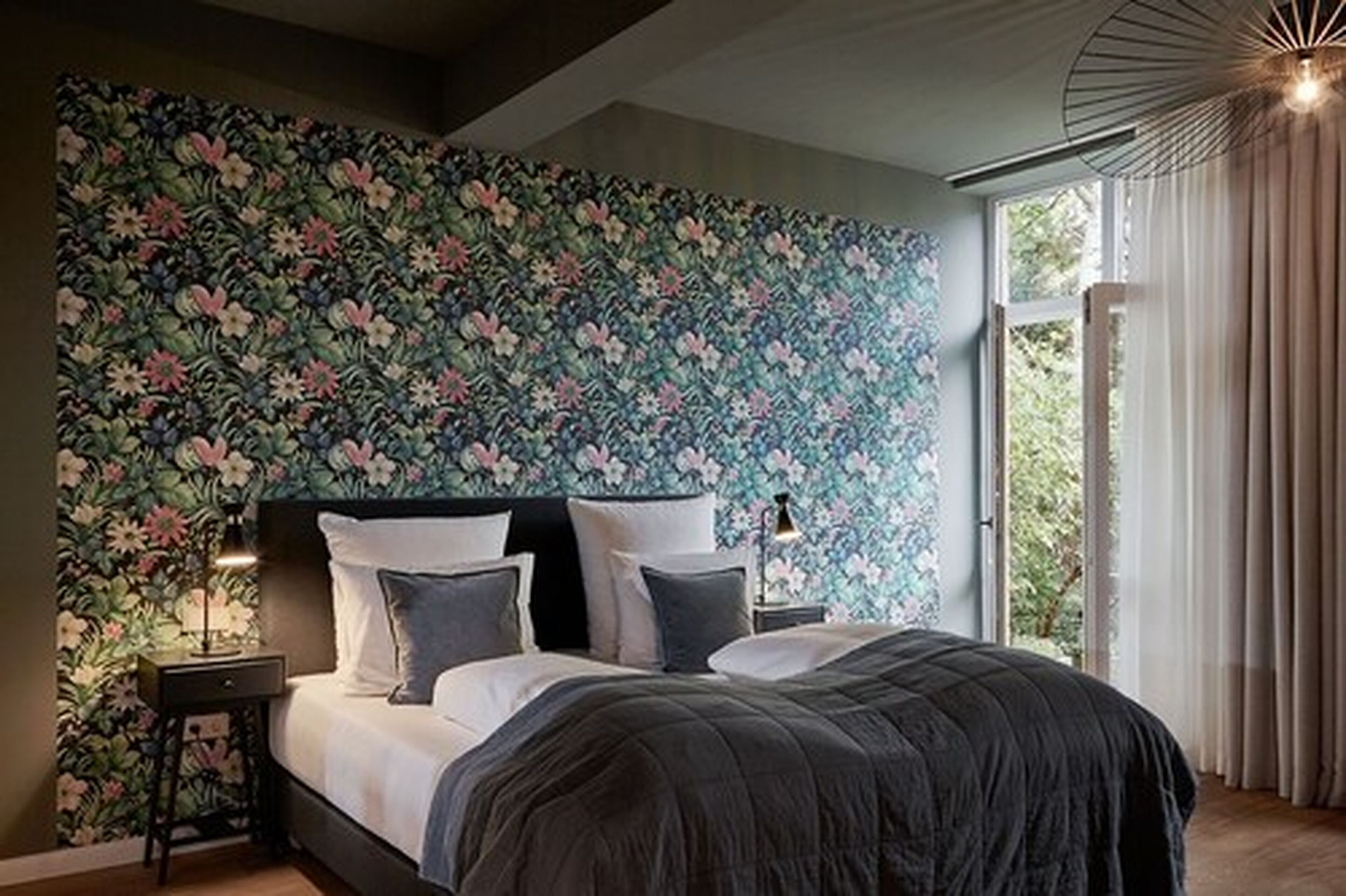 Une chambre à coucher avec un papier peint à motifs floraux, un lit avec une literie sombre, une table de chevet et une fenêtre avec des rideaux.