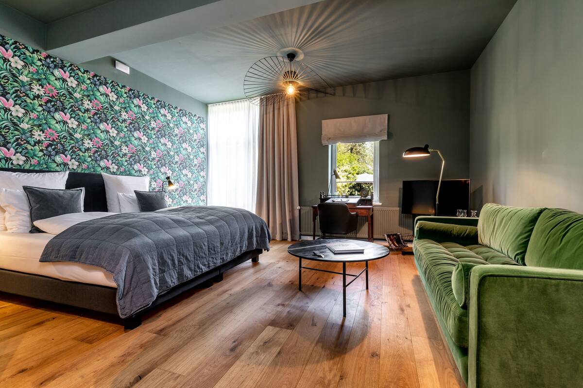 A modern bedroom with a large bed, a green sofa, a desk with a chair, and a window with curtains.