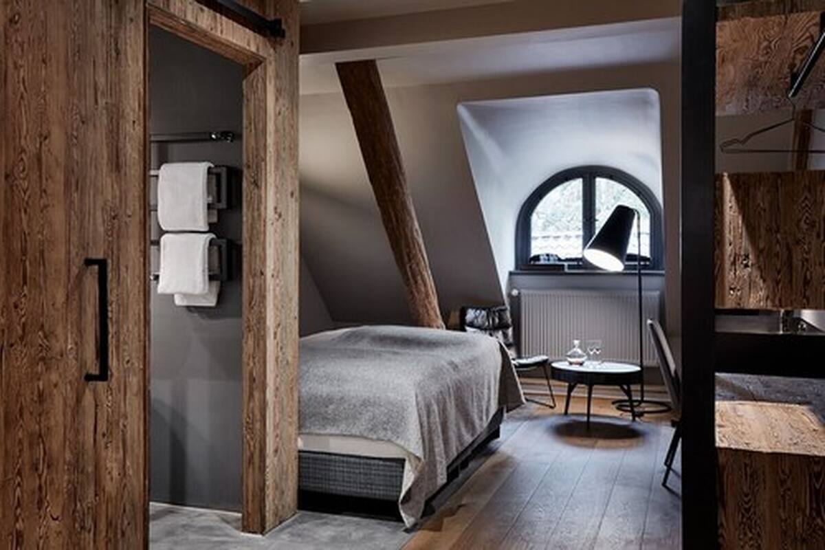 A bedroom with a sloped ceiling, a bed, a small table with a lamp, and a window with a view.