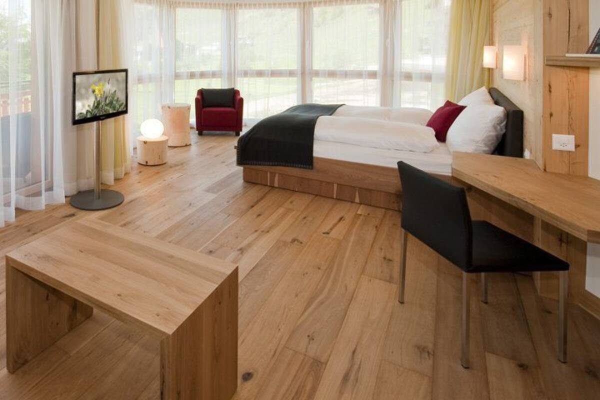 Ett modernt rum med trägolv, en säng med vitt och svart sänglinne, ett skrivbord med en stol, en tv och utsikt över naturen genom stora fönster.