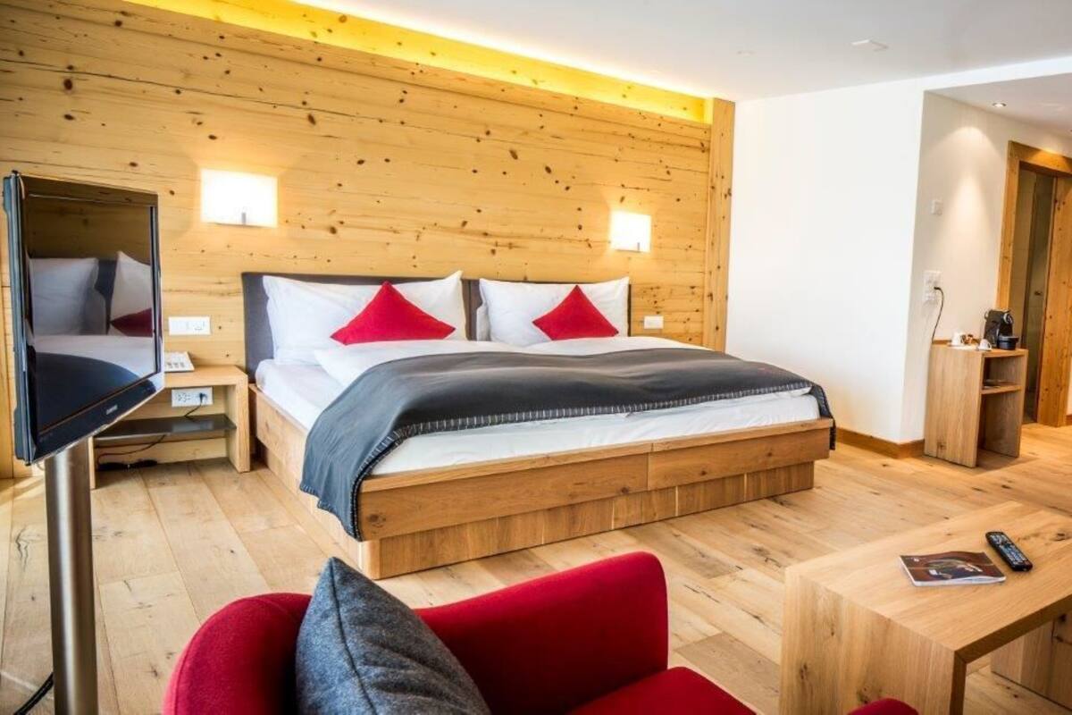 Ett modernt hotellrum med en sänggavel i trä, en stor säng med röda och vita kuddar, en tv, ett skrivbord i trä och en röd stol.