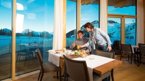 A dining area with a table set for two, large windows offering a view of a snowy landscape, and a wooden deck outside. - Hotel Royal (Riederalp)