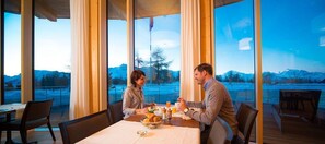 A dining area with a table set for two, large windows with a view of mountains and a lake, and a wooden deck outside. - Hotel Royal (Riederalp)