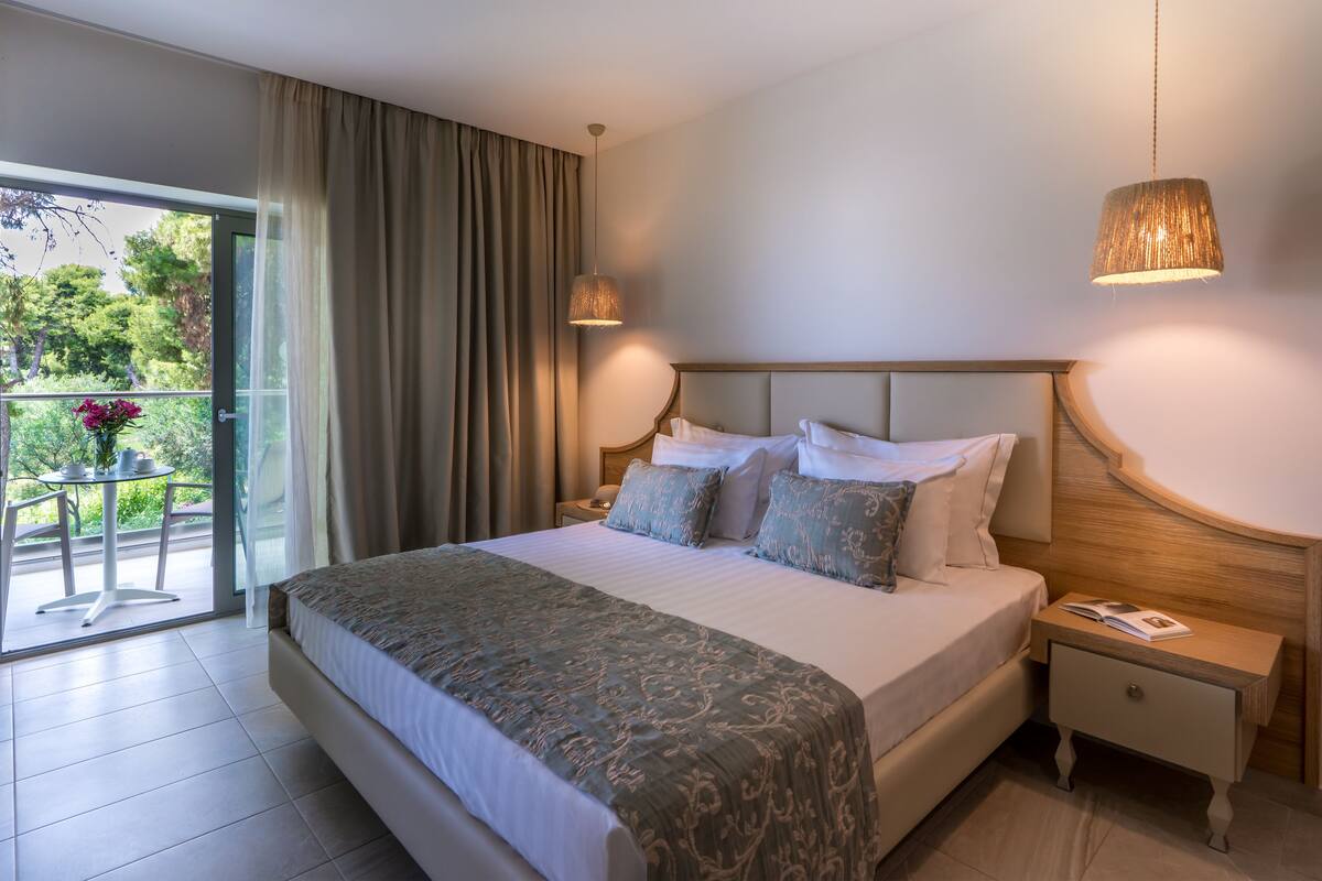 A hotel room with a large bed, a nightstand, a bedside table, and a view of a balcony with a table and chairs.