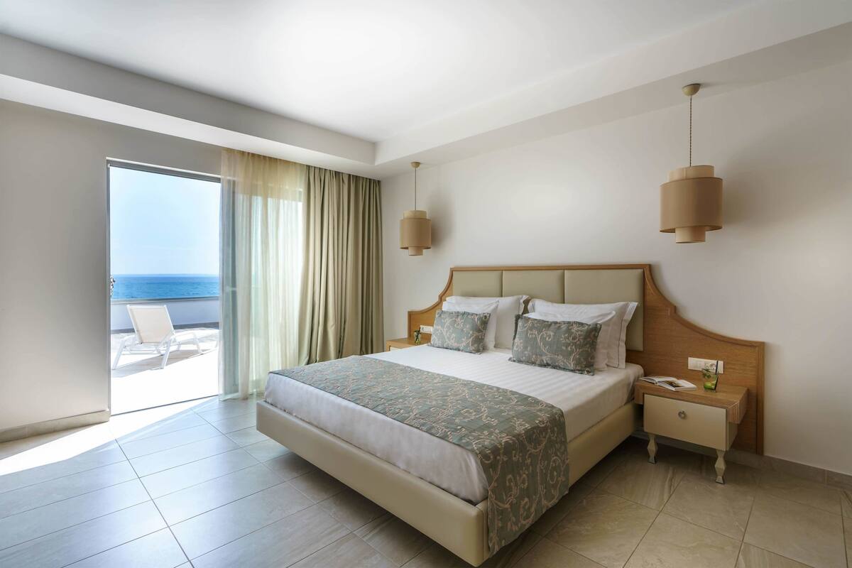A hotel room with a large bed, a nightstand, a bedside table, a window with curtains, and a view of the ocean.