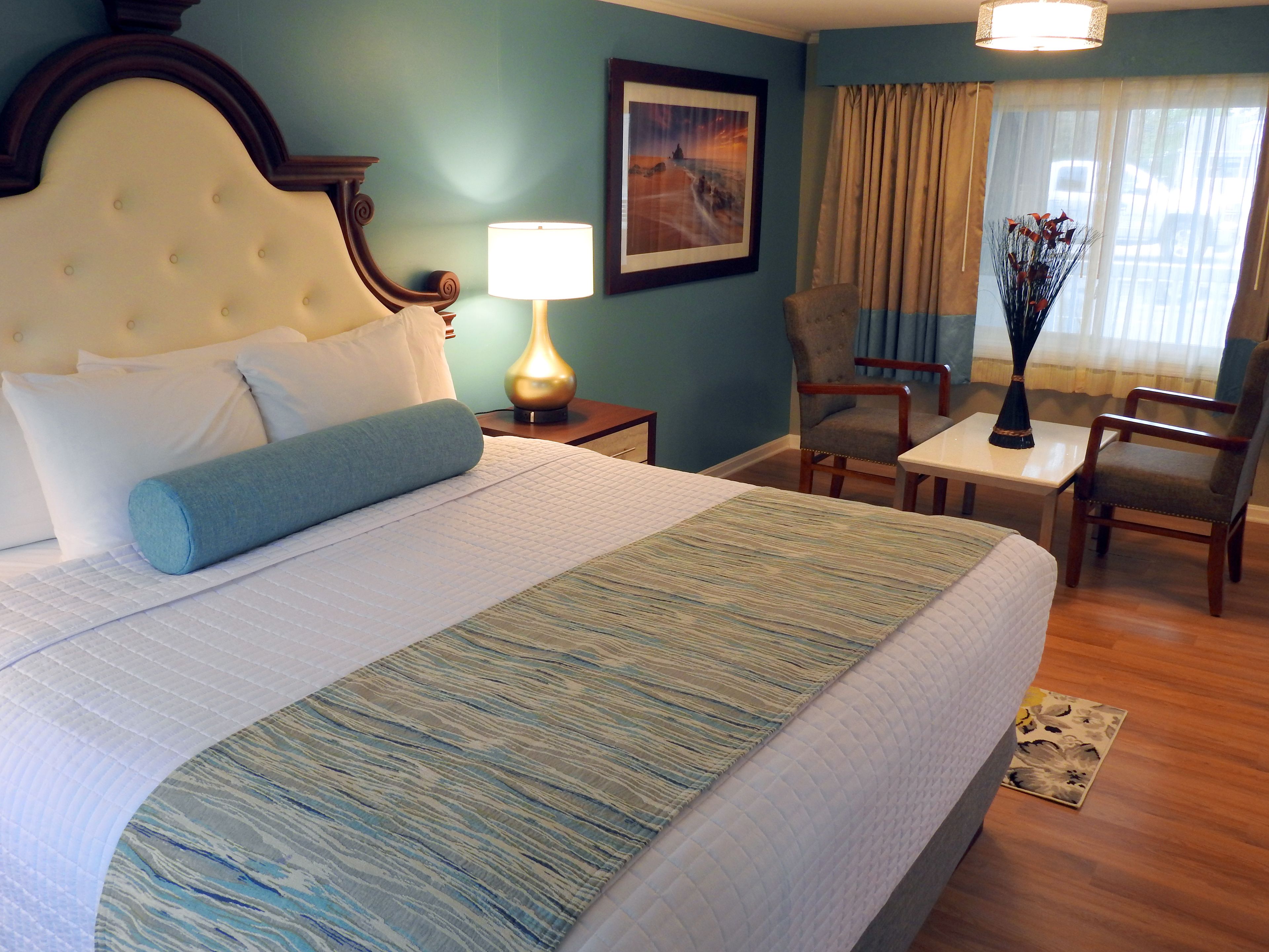 A hotel room with a large bed, a nightstand, a lamp, a chair, a table, and a painting on the wall.