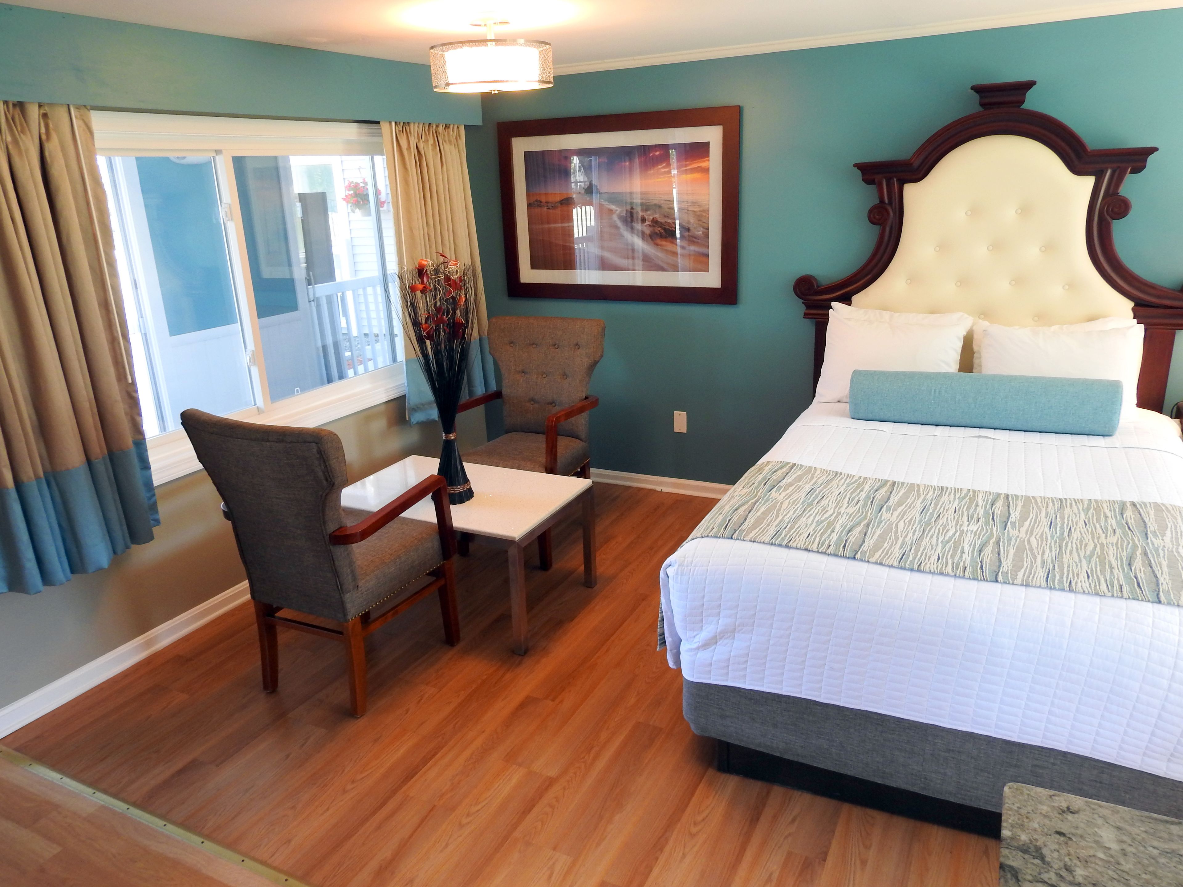 A hotel room with a bed, a chair, a small table, a vase with flowers, and a picture on the wall.