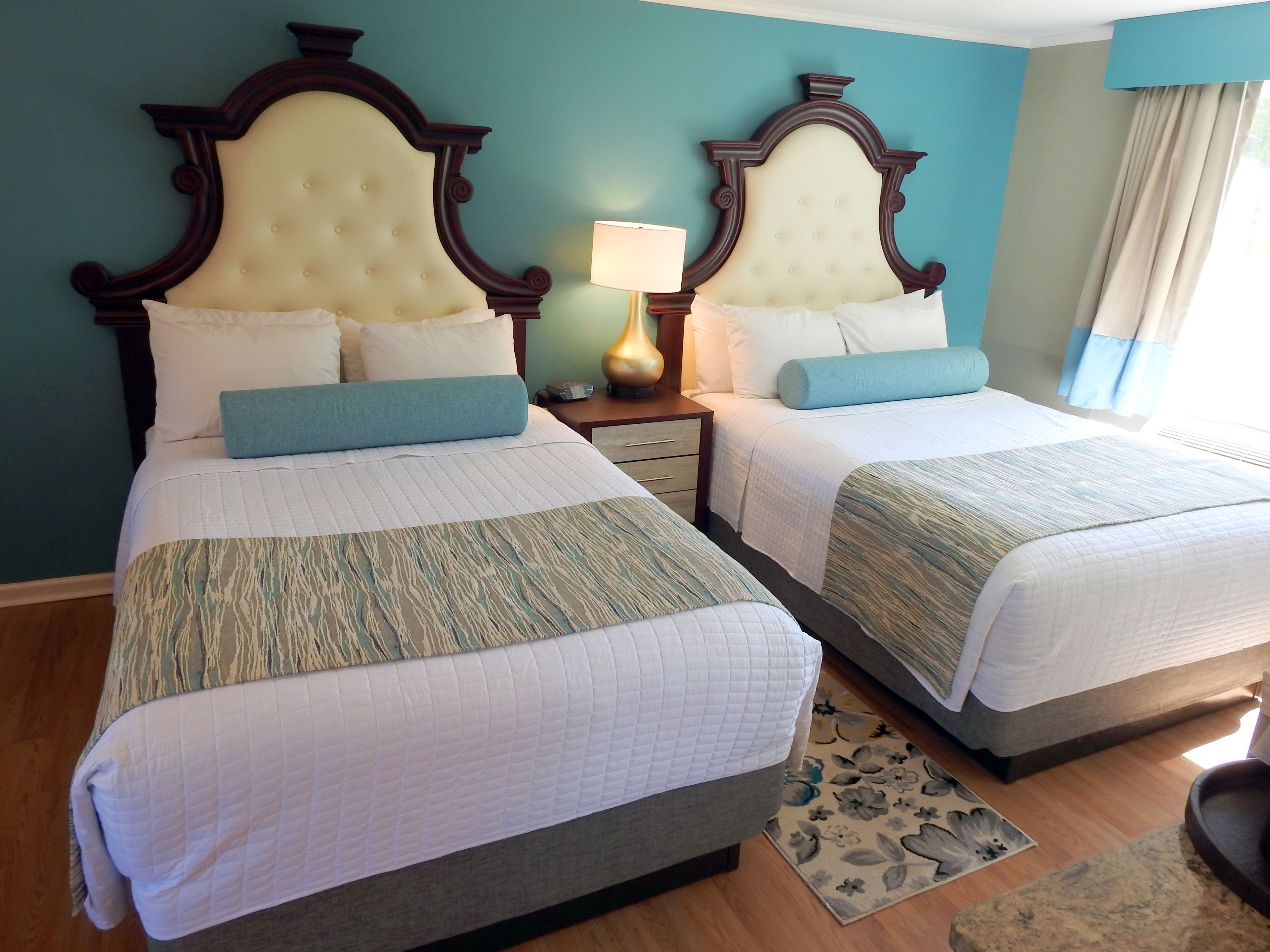 A hotel room with two queen beds, a nightstand with a lamp, and a floral rug.