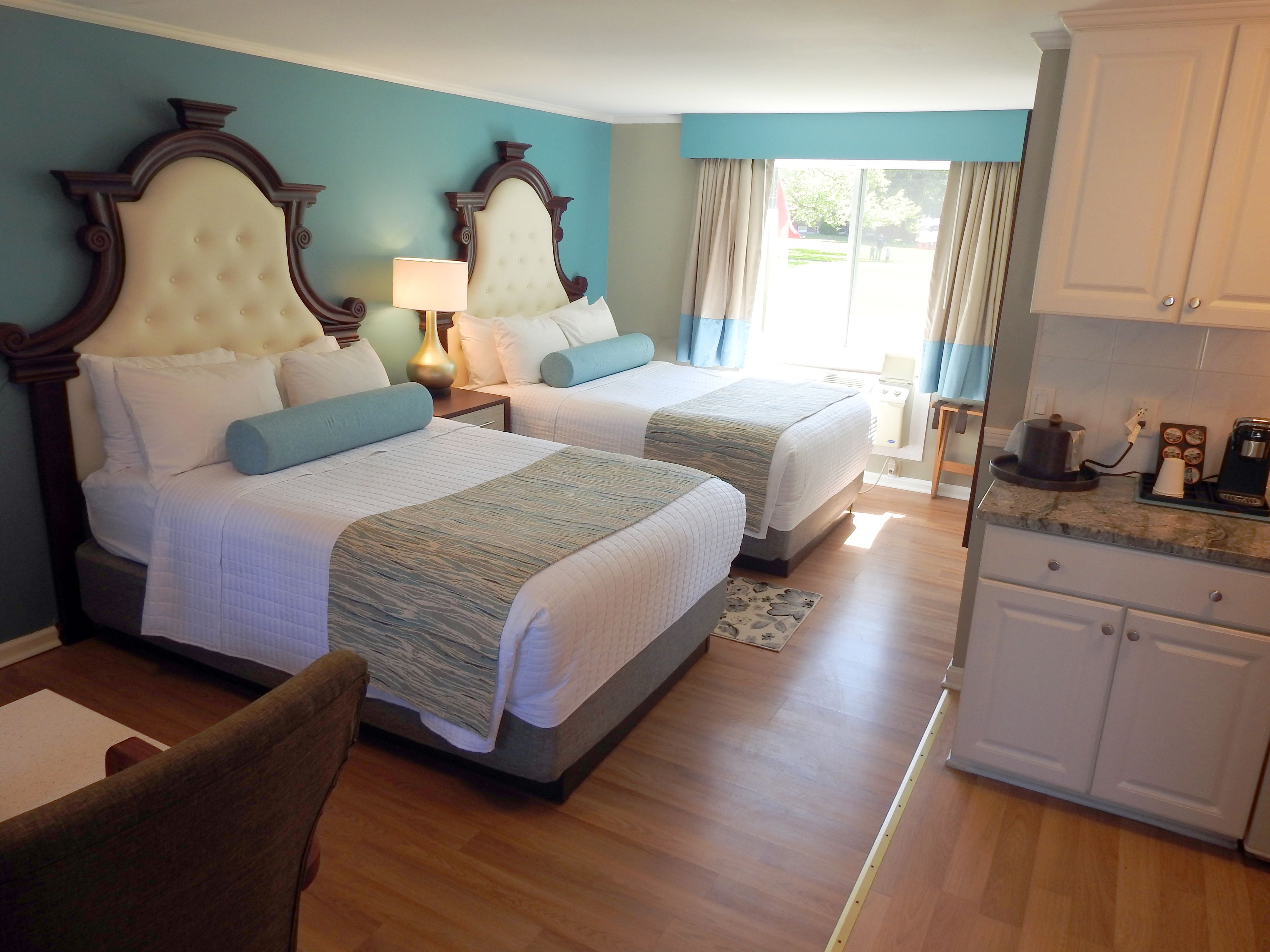 Habitación de hotel con dos camas, escritorio, silla, zona de cocina y vistas al exterior.
