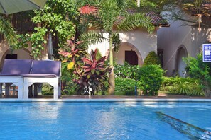 A swimming pool with lounge chairs and a covered seating area, surrounded by lush greenery and a building with arches. - Diadem Resort (Phu Quoc)