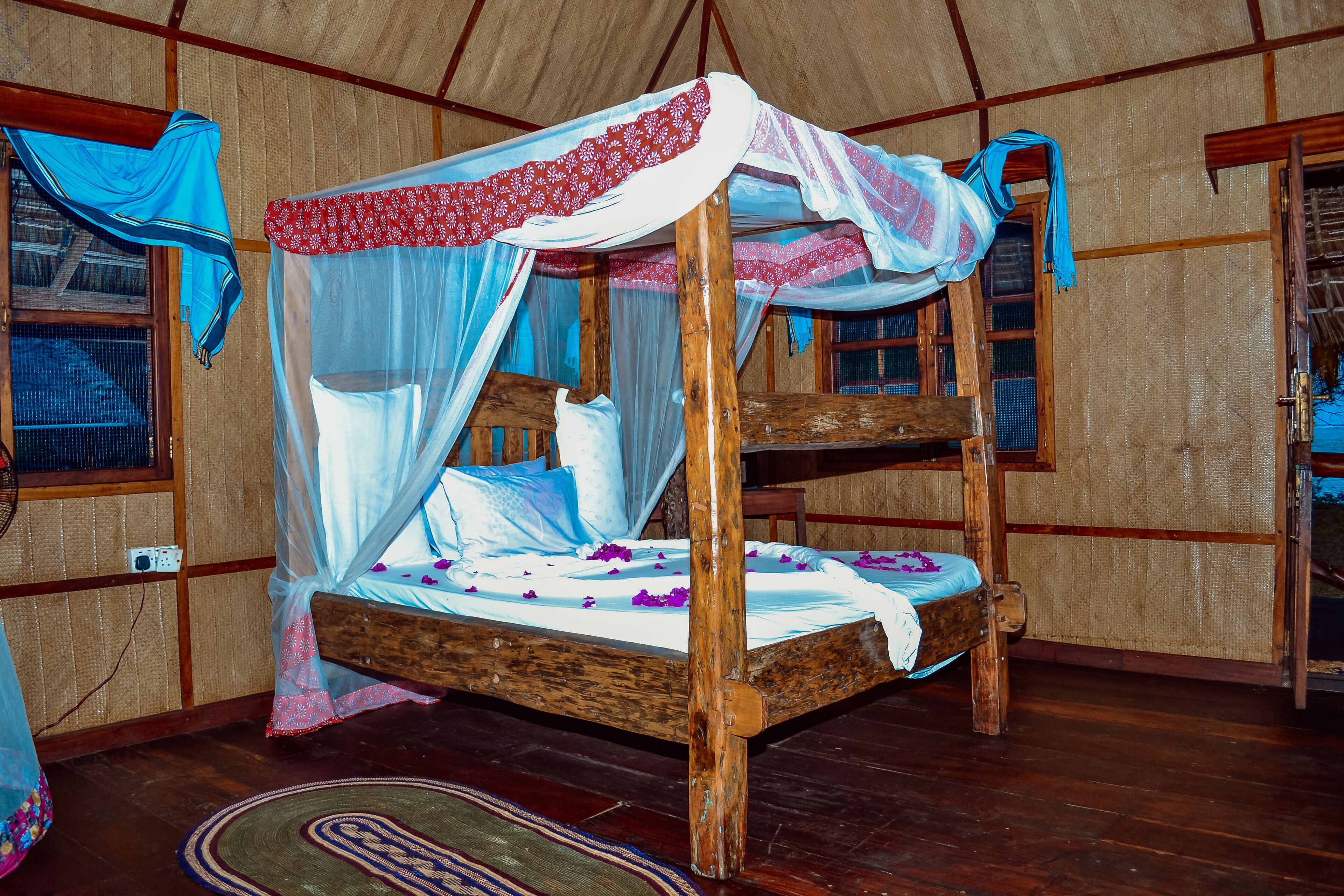 A wooden bunk bed with a canopy, a woven mat on the floor, and a window with a curtain.