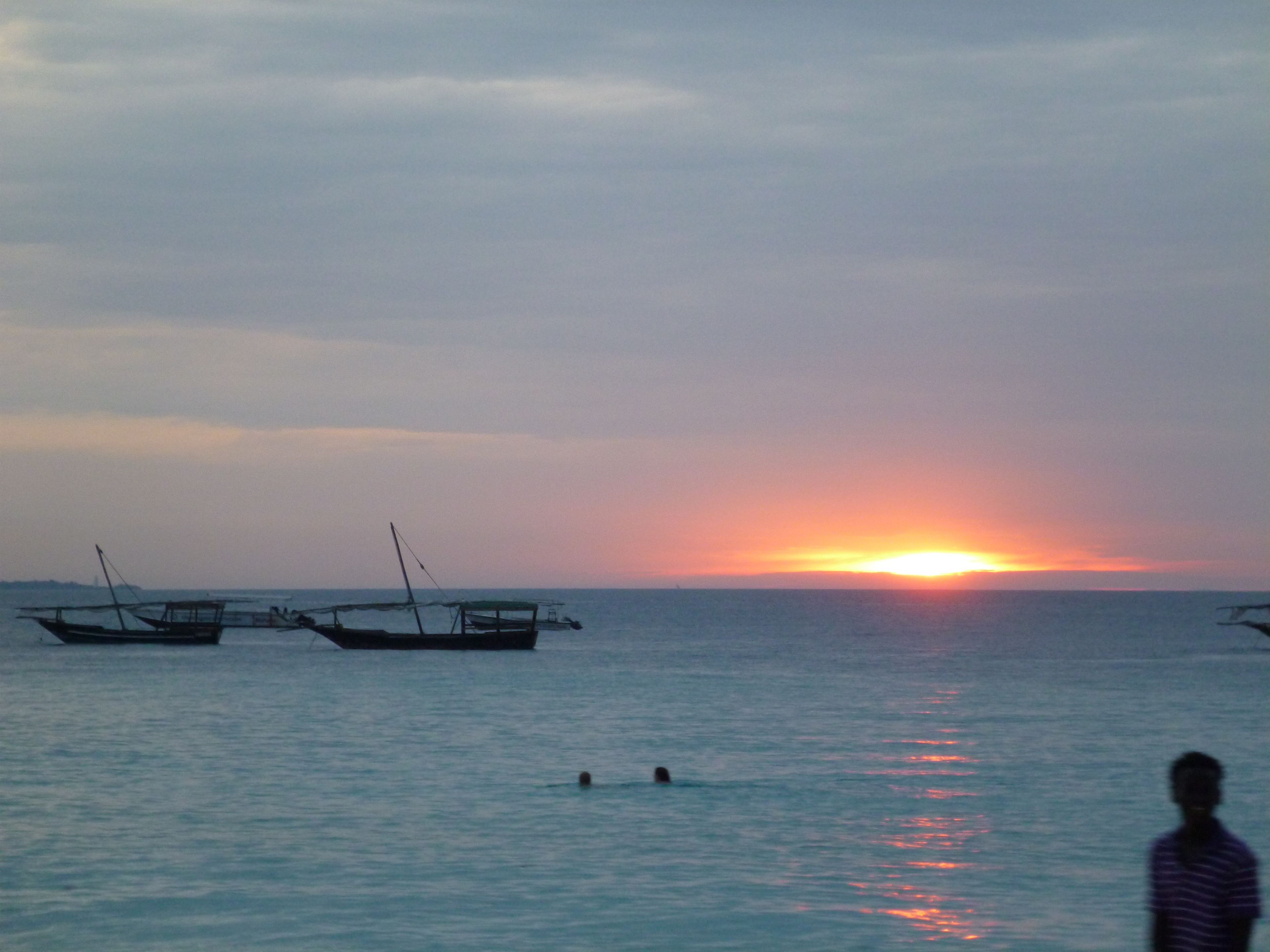A sunset over a calm sea with boats and a person swimming.