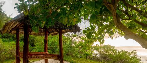 A wooden gazebo with a thatched roof, surrounded by sand and greenery, overlooking a beach.