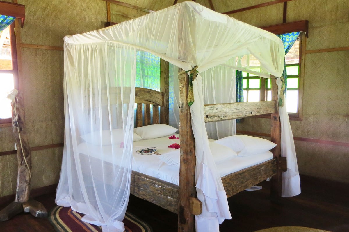 A four-poster bed with a white canopy, wooden headboard, and a woven mat on the floor.