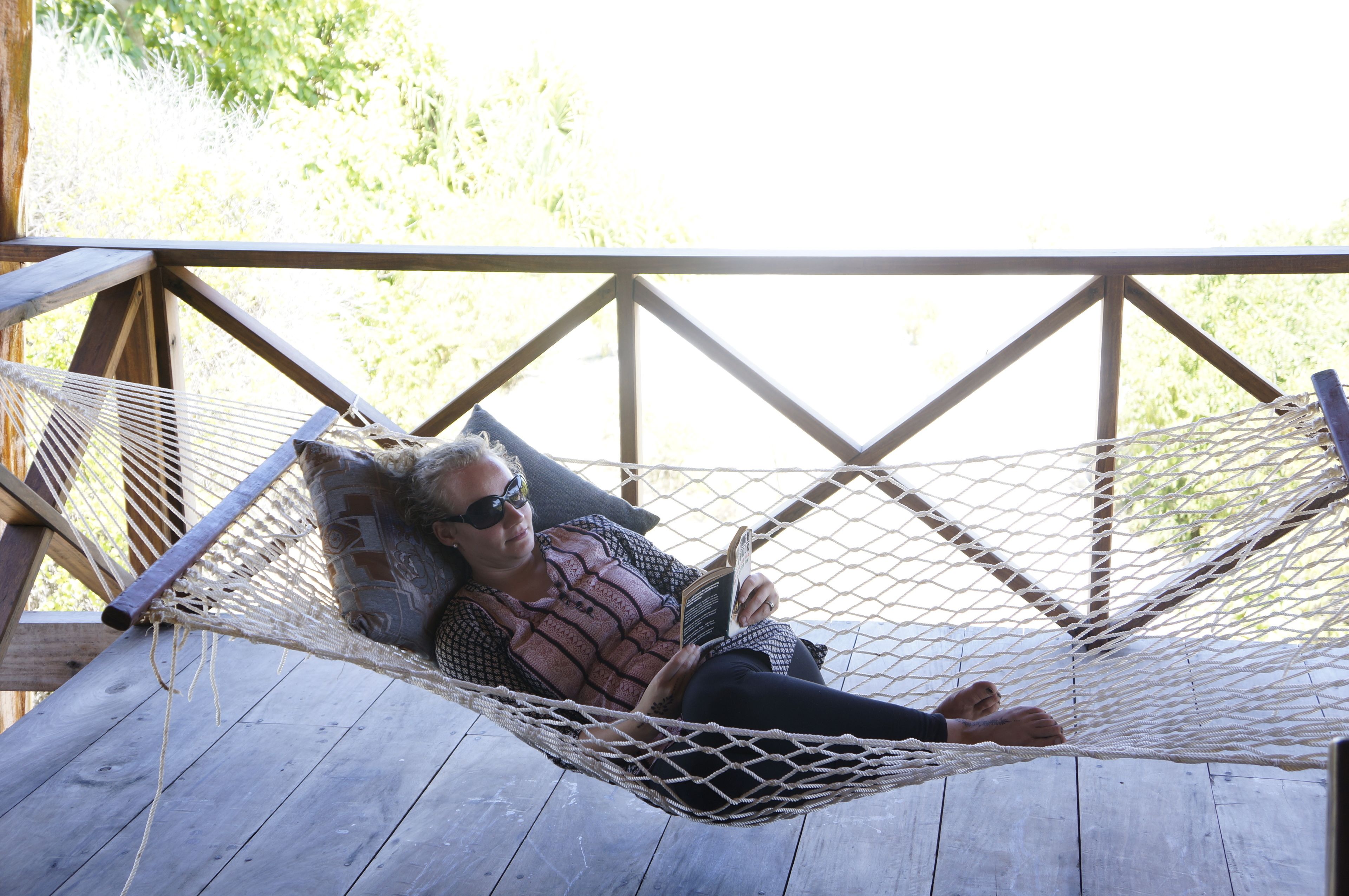 A person lying in a hammock on a wooden deck with a view of greenery in the background.