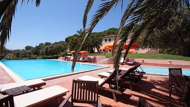 Un área de piscina con sillones de descanso y sombrillas, rodeada de vegetación y un edificio al fondo.
