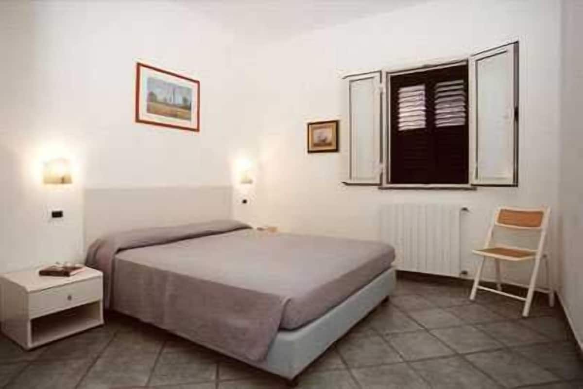 Quarto com cama, mesas de cabeceira, uma cadeira e janela com persianas.