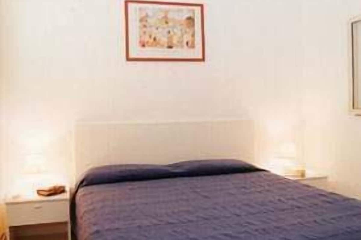 Quarto com cama, mesa de cabeceira, um quadro na parede e piso ladrilhado.