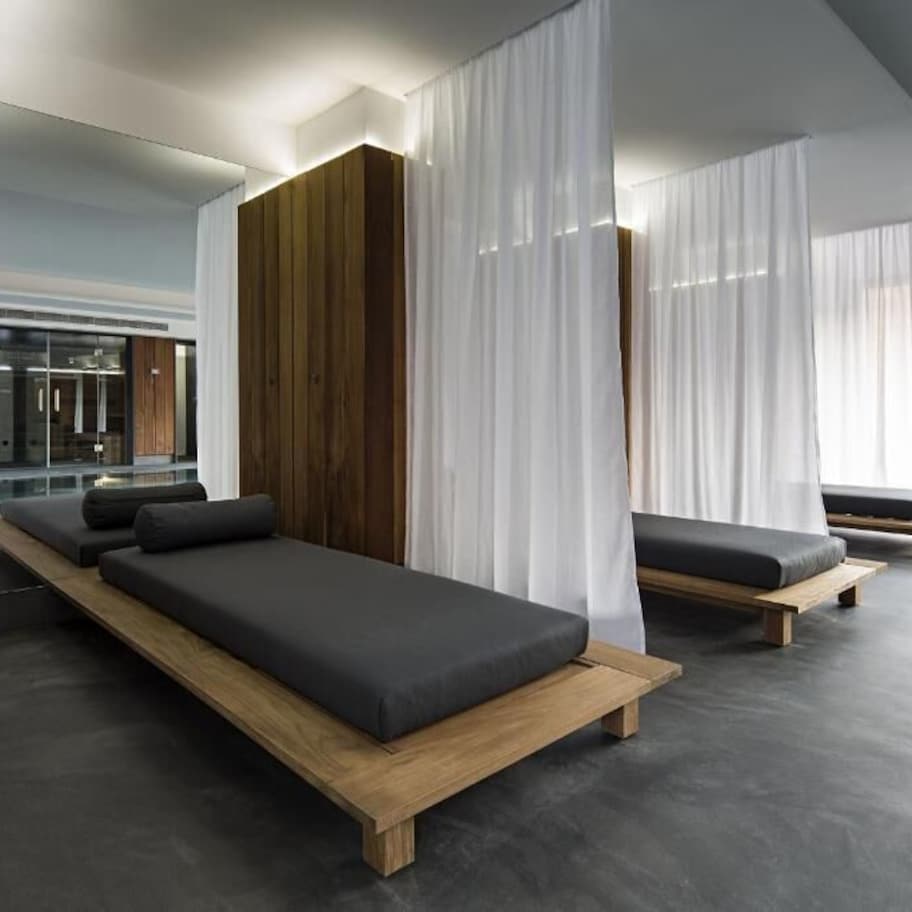A modern spa room with two wooden beds, white curtains, and a large window offering a view of the outdoors.