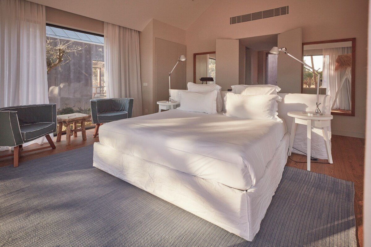 A modern hotel room with a large bed, a desk, a chair, and a small table.