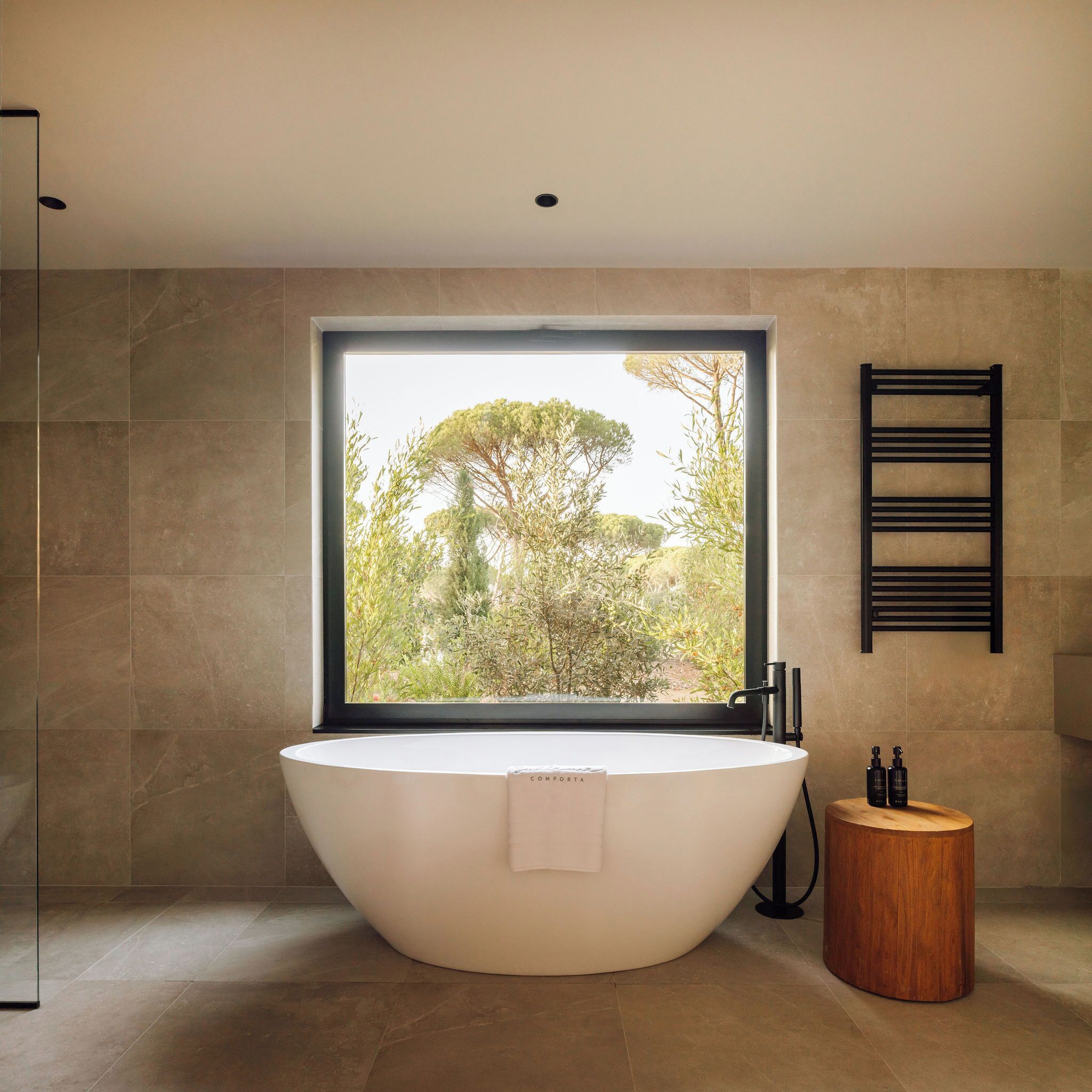 A modern bathroom with a freestanding bathtub, a window with a view of trees, and a wooden stool.
