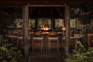 A bar area with a fire feature, wooden chairs, and a view through large windows.