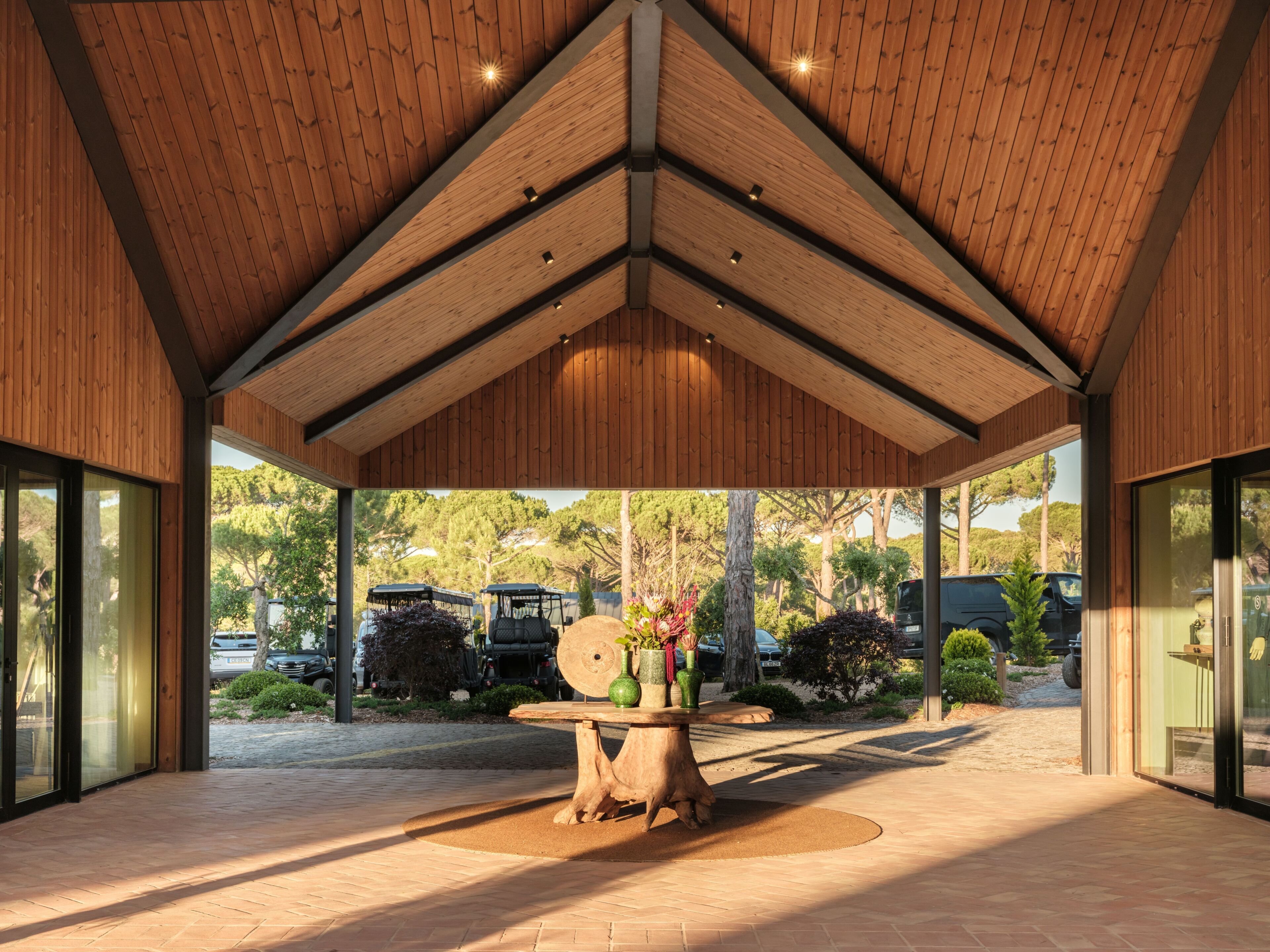 A covered outdoor area with a wooden ceiling, a table with vases, and a view of parked cars and trees.