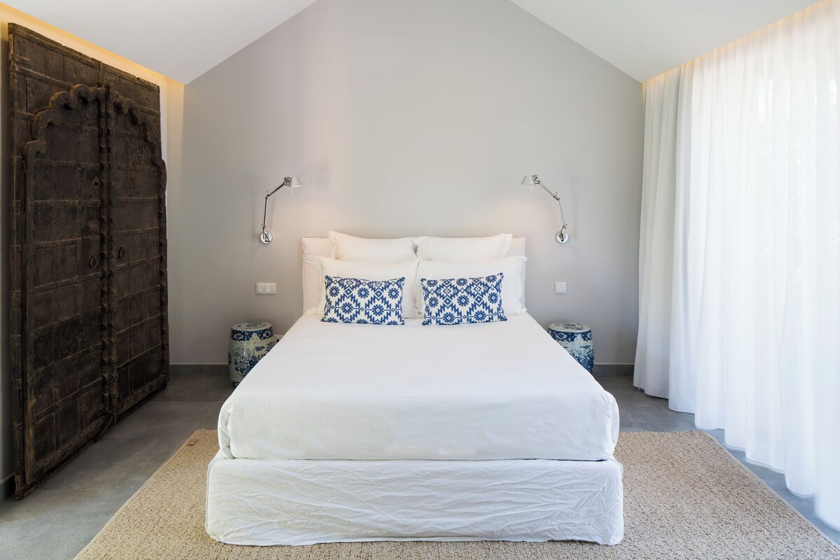 A bedroom with a bed, two bedside lamps, a large window with curtains, and a wooden door.