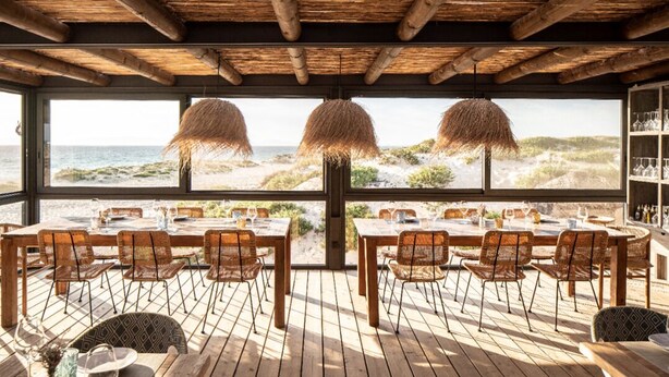 Un área de comedor al aire libre con mesas y sillas de madera, ventanas amplias con vista a la playa y al océano, y sombrillas de paja.