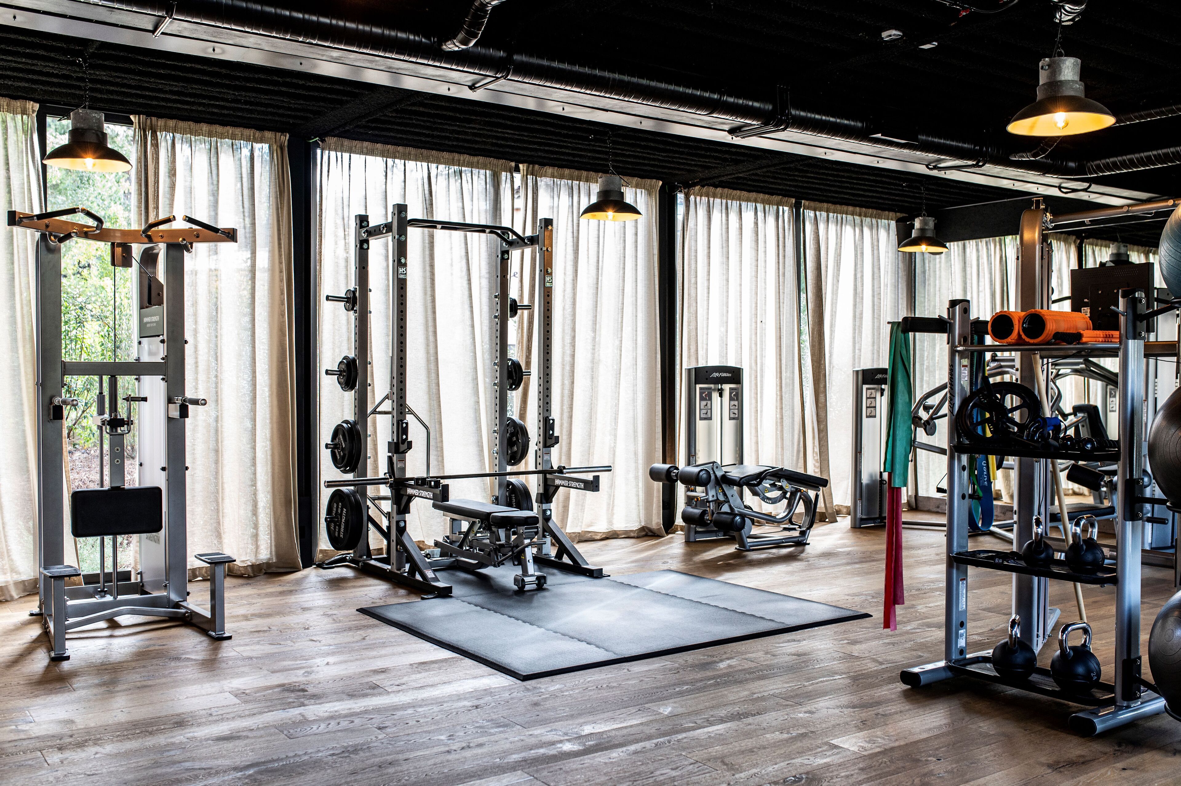 Un gimnasio con diferentes equipos de ejercicio, ventanas amplias con cortinas y suelo de madera.