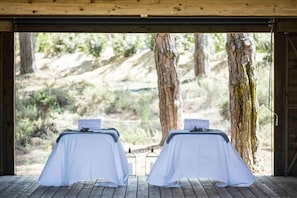 A room with two beds covered in white sheets, a wooden ceiling, and a view of a rocky outdoor landscape.