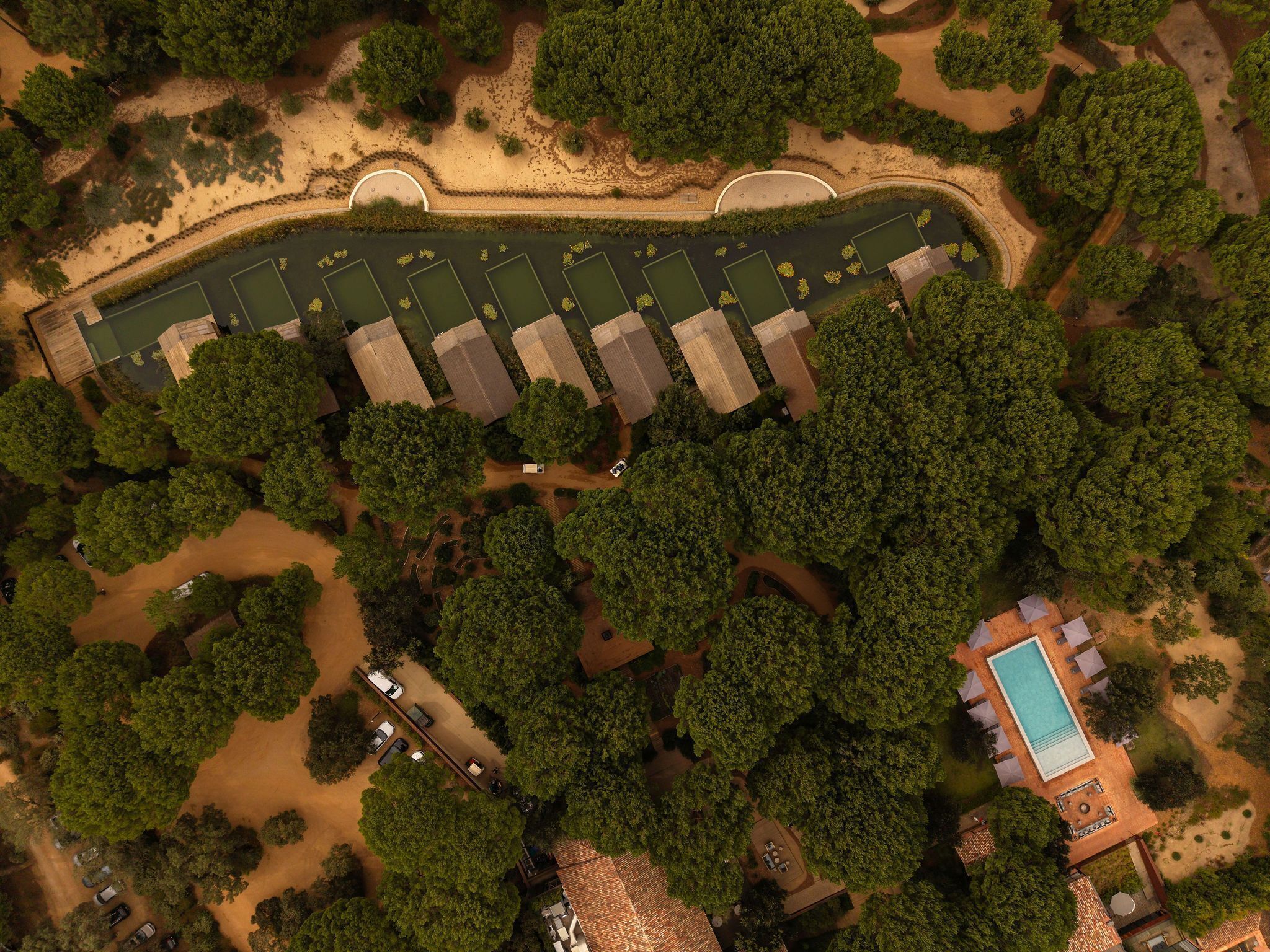 An aerial view of a resort with multiple buildings, a swimming pool, and a parking area surrounded by dense trees.