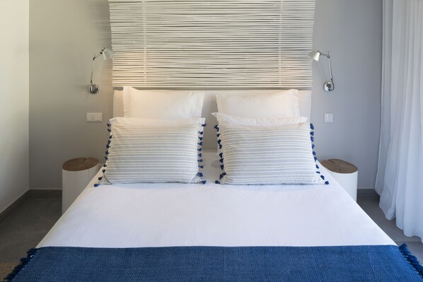 A neatly made bed with white bedding and blue accents, two bedside lamps, and a window with blinds. - Sublime Comporta Country Retreat & SPA (Grandola)