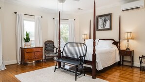 Une chambre à coucher avec un lit à baldaquin, une table de chevet avec une lampe, une commode en bois, une chaise et une grande fenêtre avec des rideaux.