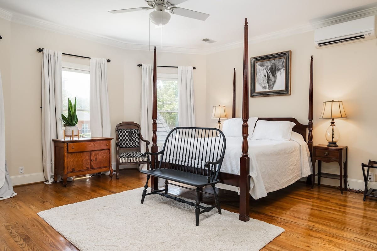 A bedroom with a four-poster bed, a nightstand with a lamp, a wooden dresser, a chair, and a large window with curtains.