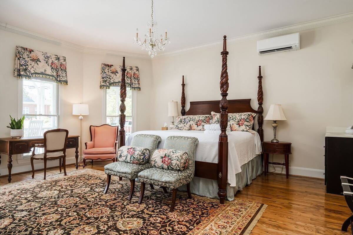 A bedroom with a four-poster bed, a chandelier, a desk with a chair, a small table with a lamp, and a large ornate rug.