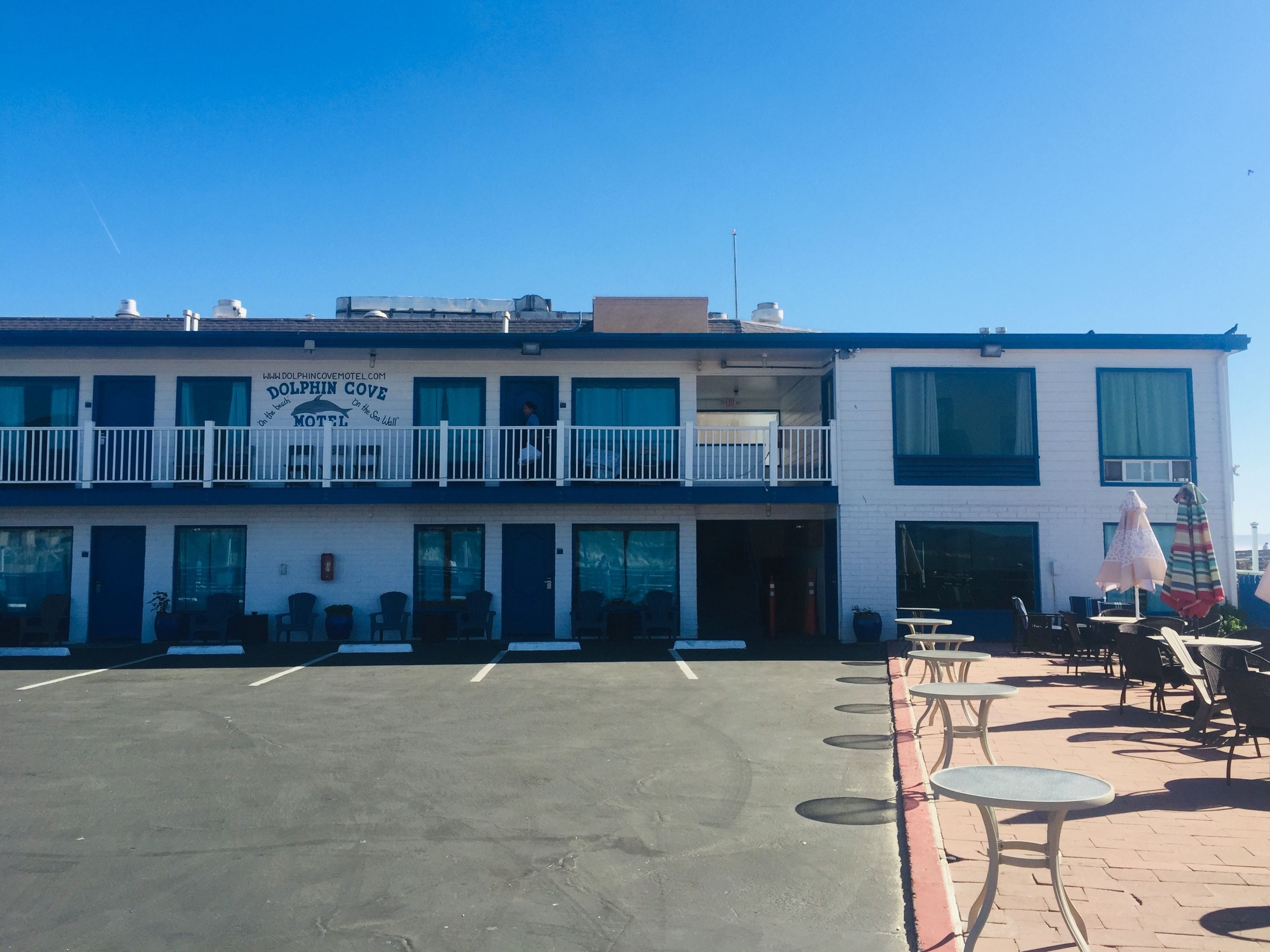 A two-story motel with a balcony on the upper floor, outdoor seating, and a clear sky.