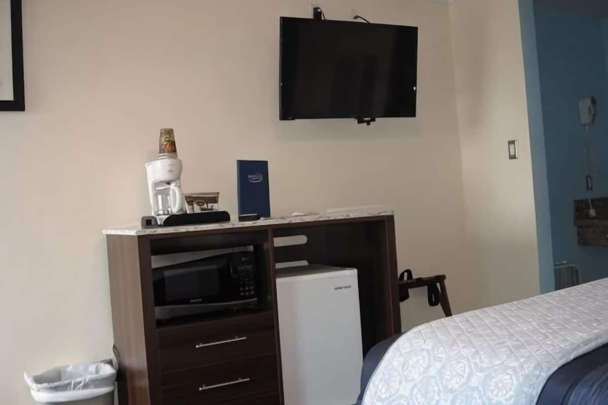 Een slaapkamer met een bed, een bureau met een magnetron, een koffiezetapparaat, een aan de muur bevestigde televisie en een prullenbak.
