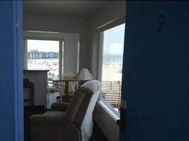 A room with a view of the beach, a sofa, a table with a lamp, and a window with blinds.