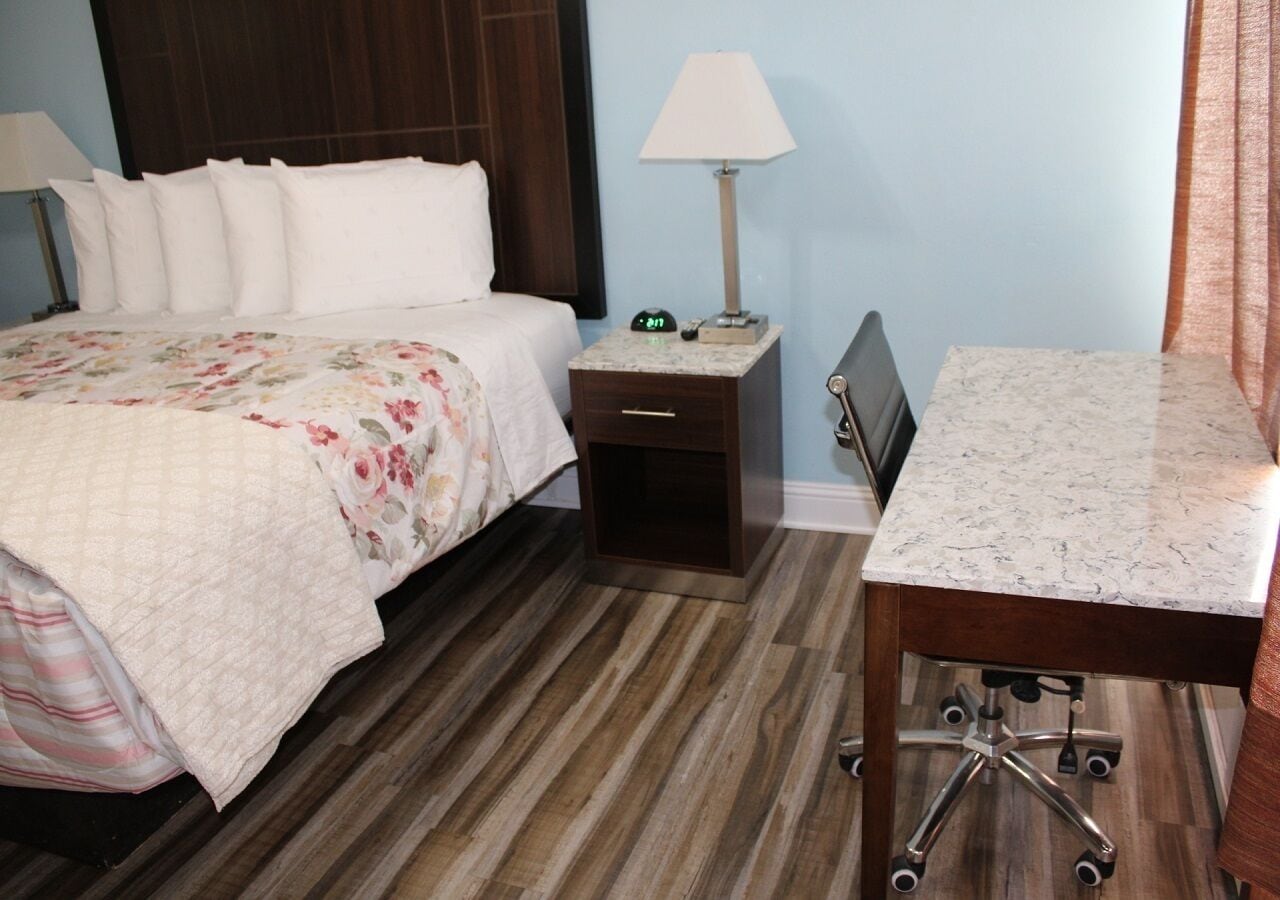 Une chambre d’hôtel avec un lit, une table de chevet avec une lampe, un bureau avec une chaise et un plancher en bois.
