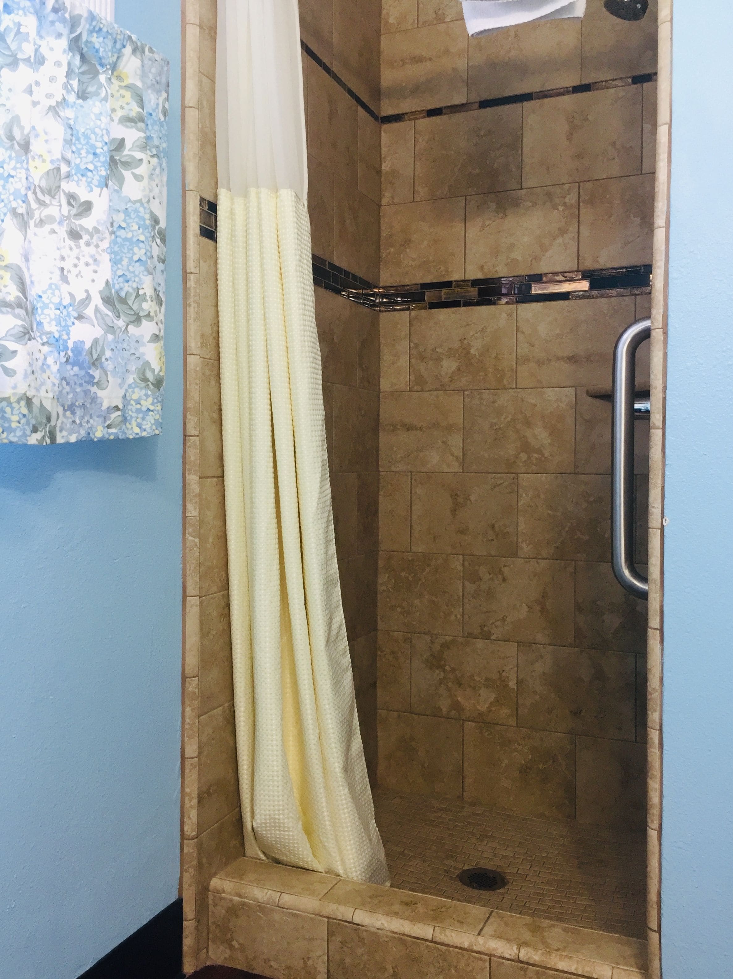 A shower area with a beige tiled wall, a white shower curtain, and a metal grab bar.