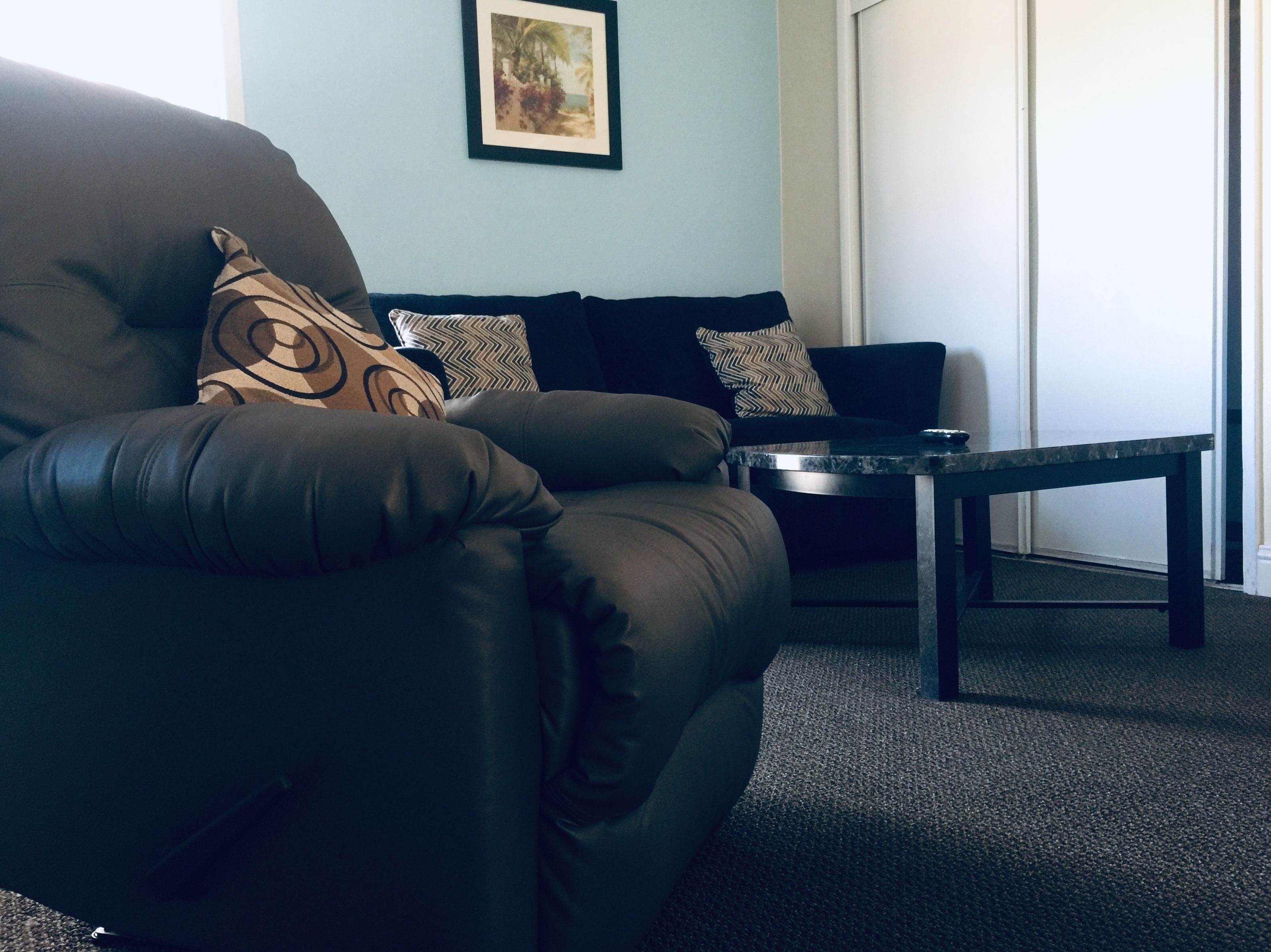 A dark brown leather sofa with patterned cushions, a small side table, and a framed picture on the wall.