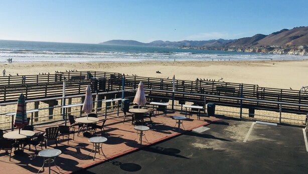 Une terrasse au bord de la plage, avec des tables et des chaises, d’où l’on a une vue sur l’océan et les collines.